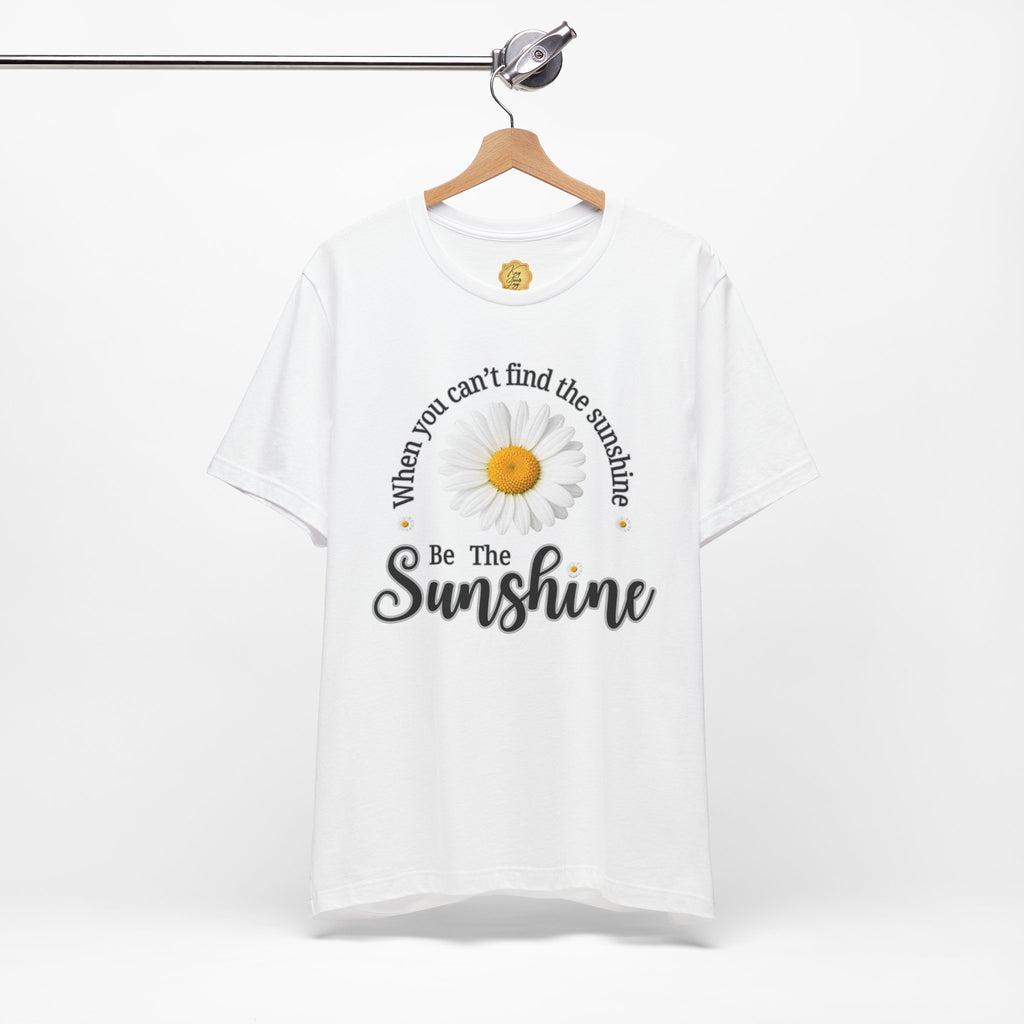 A white short-sleeve t-shirt hanging on a wooden hanger against a light gray background. The shirt features a graphic print of a daisy with the text 'When you can't find the sunshine Be The Sunshine' in black and white lettering.