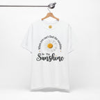 A white short-sleeve t-shirt hanging on a wooden hanger against a light gray background. The shirt features a graphic print of a daisy with the text 'When you can't find the sunshine Be The Sunshine' in black and white lettering.