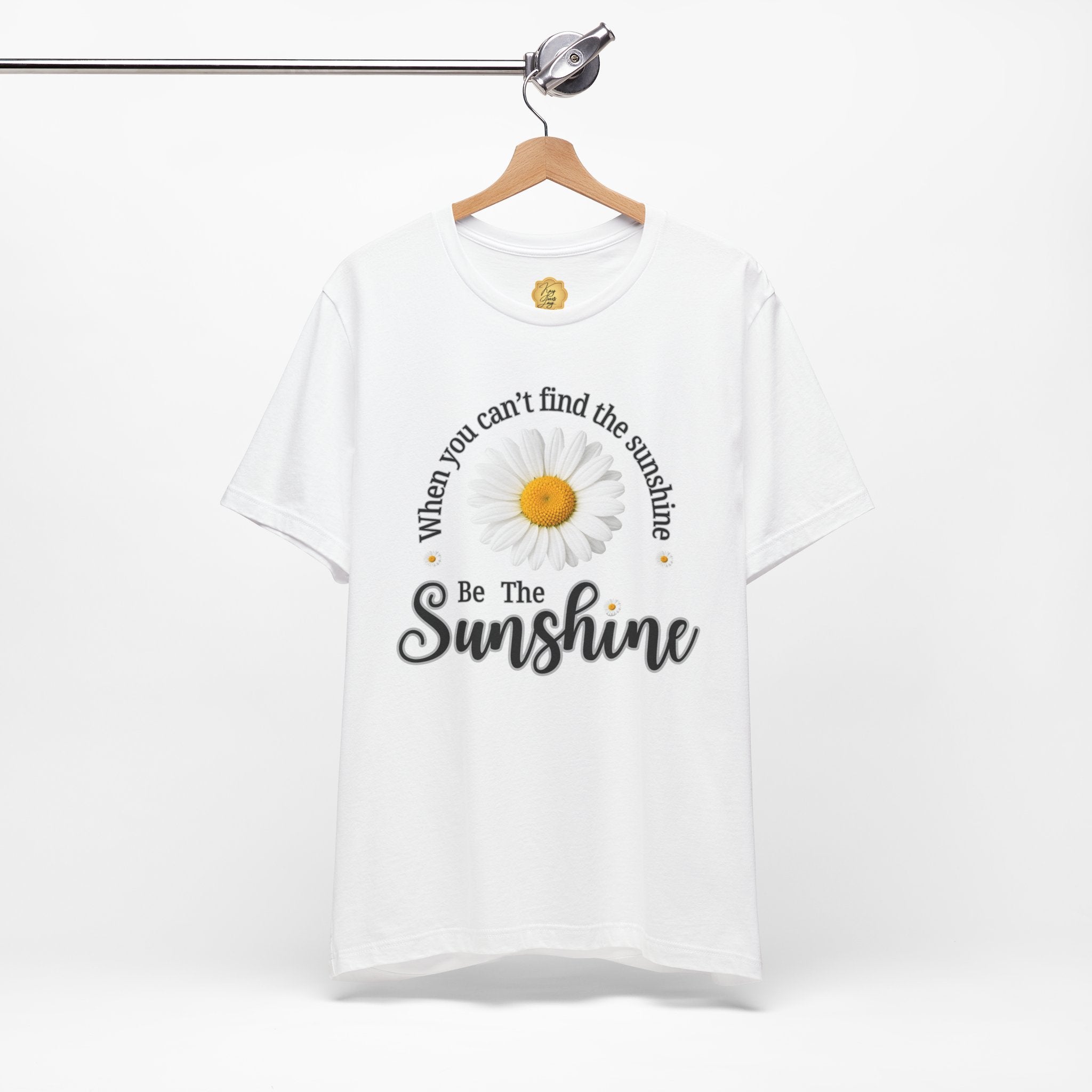 A white short-sleeve t-shirt hanging on a wooden hanger against a light gray background. The shirt features a graphic print of a daisy with the text 'When you can't find the sunshine Be The Sunshine' in black and white lettering.