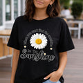A woman wearing a black graphic t-shirt featuring a large white daisy with a yellow center and the text 'When you can't find the sunshine Be The Sunshine' in white script.