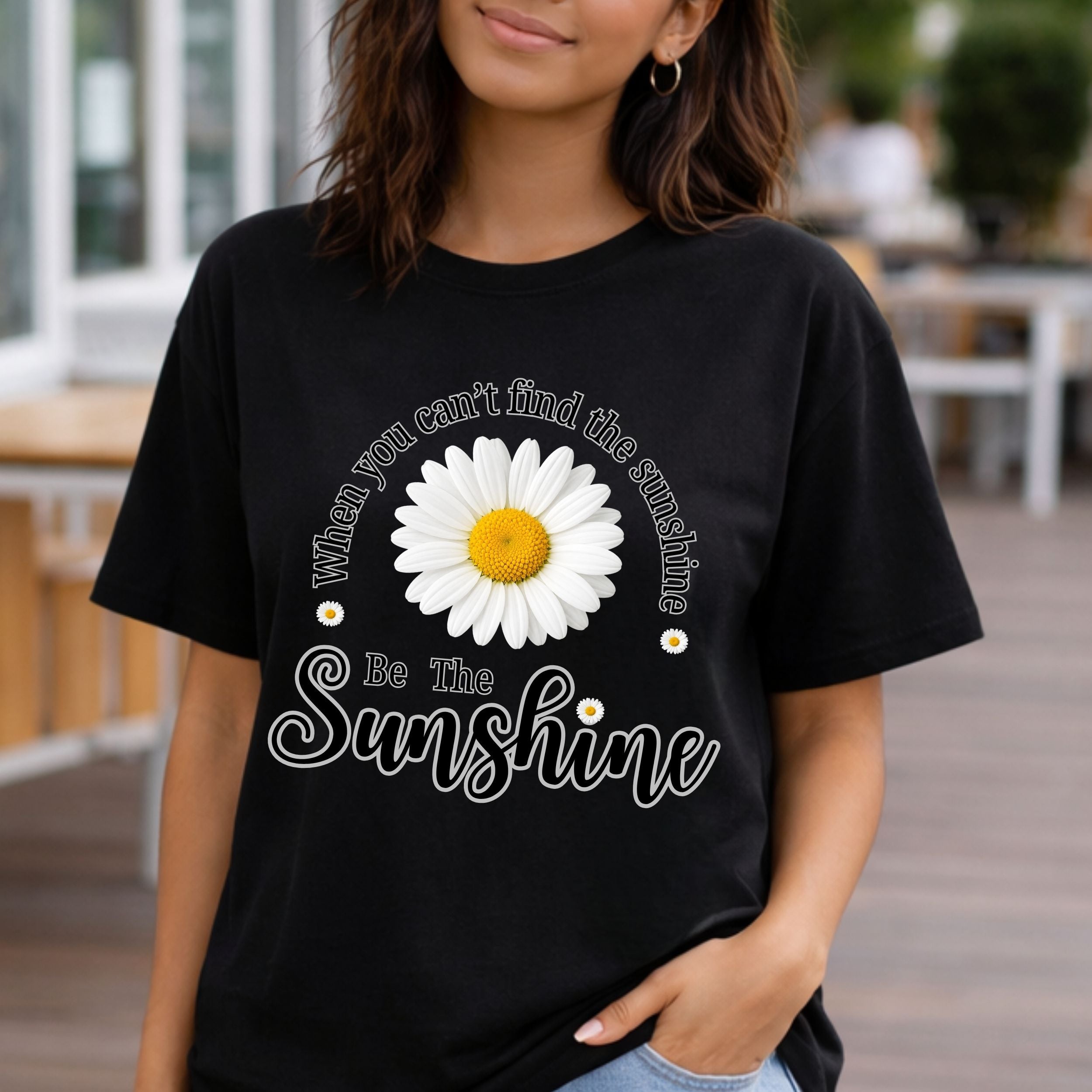A woman wearing a black graphic t-shirt featuring a large white daisy with a yellow center and the text 'When you can't find the sunshine Be The Sunshine' in white script.