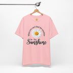 A light pink short-sleeve t-shirt hanging on a wooden hanger against a white background. The shirt features a graphic print of a white daisy with a yellow center, surrounded by the text 'When you can't find the sunshine Be The Sunshine' in black script.