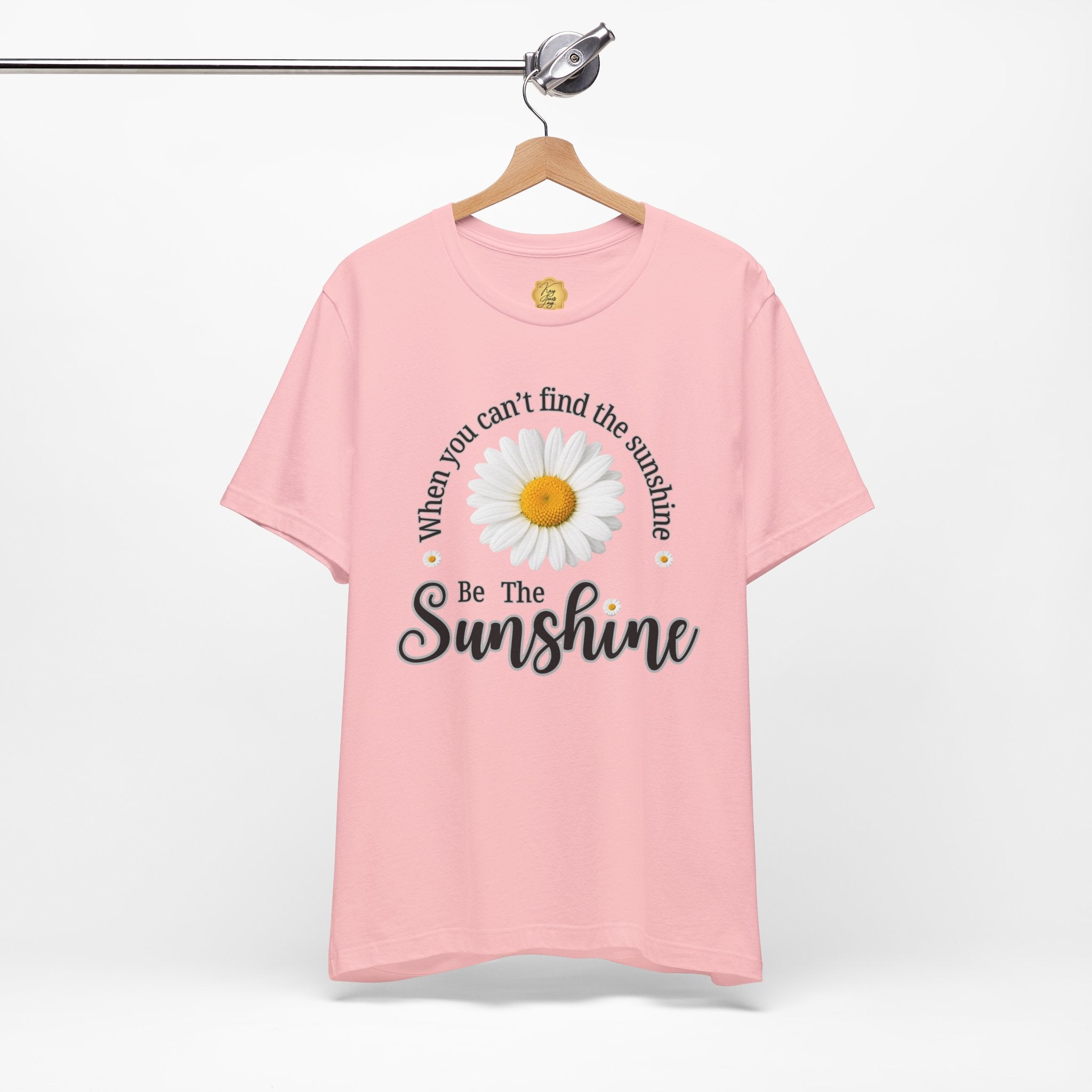 A light pink short-sleeve t-shirt hanging on a wooden hanger against a white background. The shirt features a graphic print of a white daisy with a yellow center, surrounded by the text 'When you can't find the sunshine Be The Sunshine' in black script.